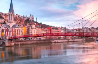 Vue emblématique de Lyon au coucher du soleil avec la passerelle rouge Saint-Georges sur la Saône, l’église Saint-Georges et la basilique de Fourvière en arrière-plan.