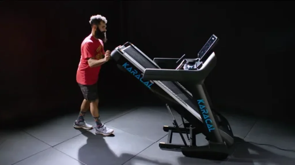 Tapis de course Moovyoo Karacal plié avec système de rangement assisté et roulettes de transport.