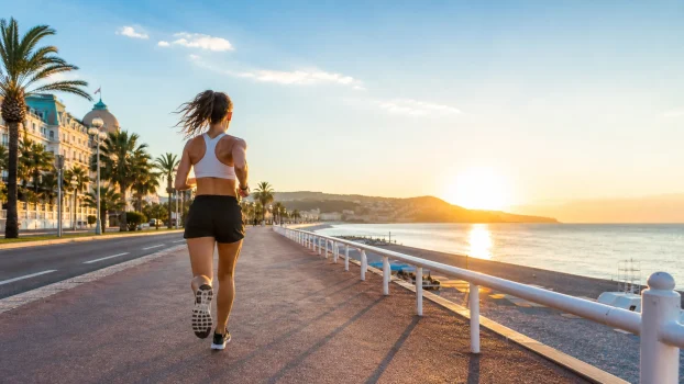 Coureuse sur la Promenade des Anglais à Nice au lever du soleil, avec la mer Méditerranée et les palmiers en arrière-plan.